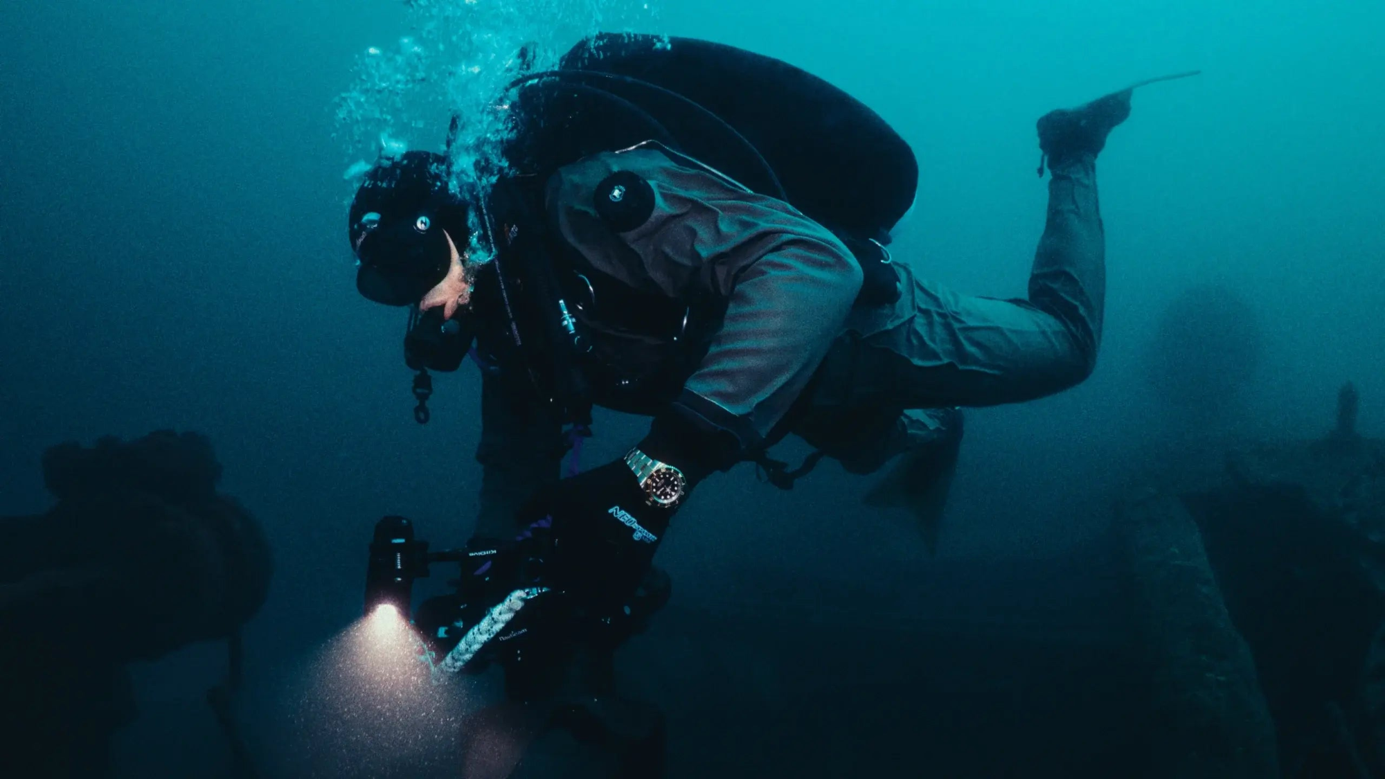 Diver underwater with diving equipment in a dark blue ocean wearing a Rolex Submariner