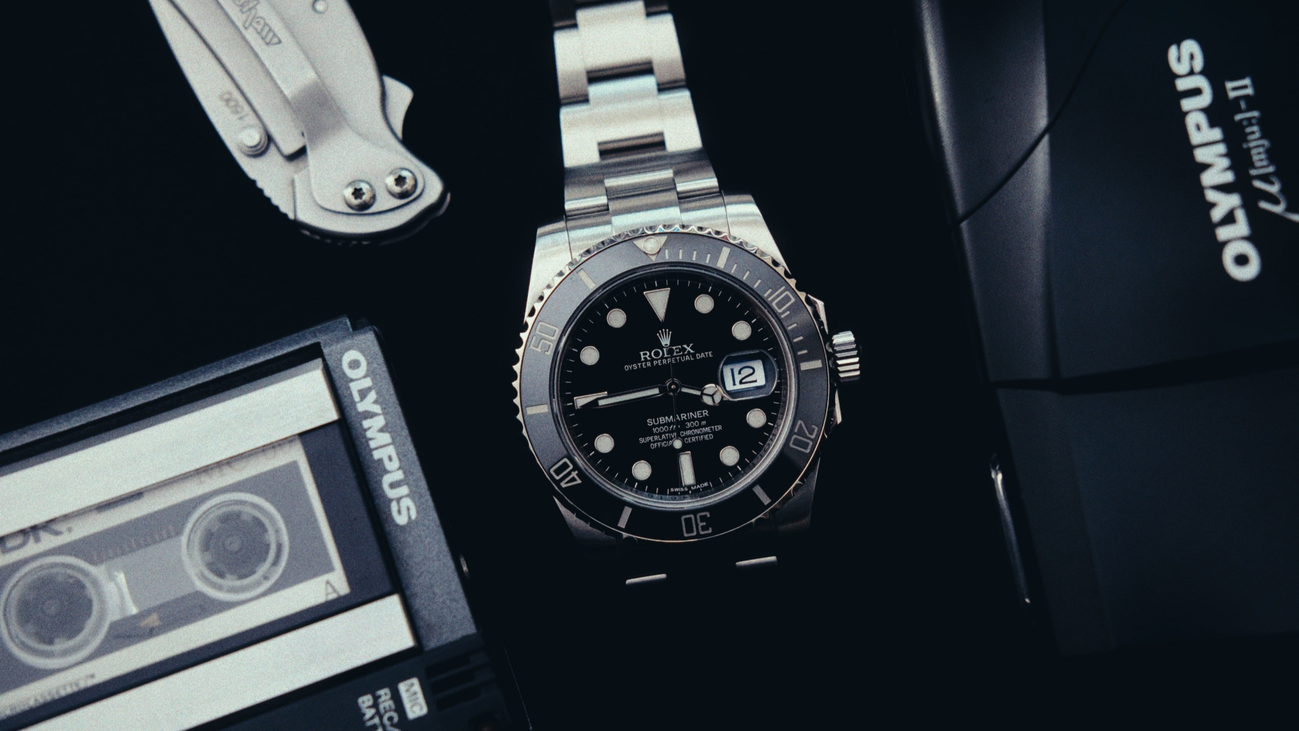 Rolex Submariner watch with black dial on a dark surface with Olympus products.